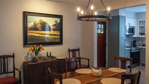 A dining room with a wooden table, chairs, a painting, and a kitchen in the background.