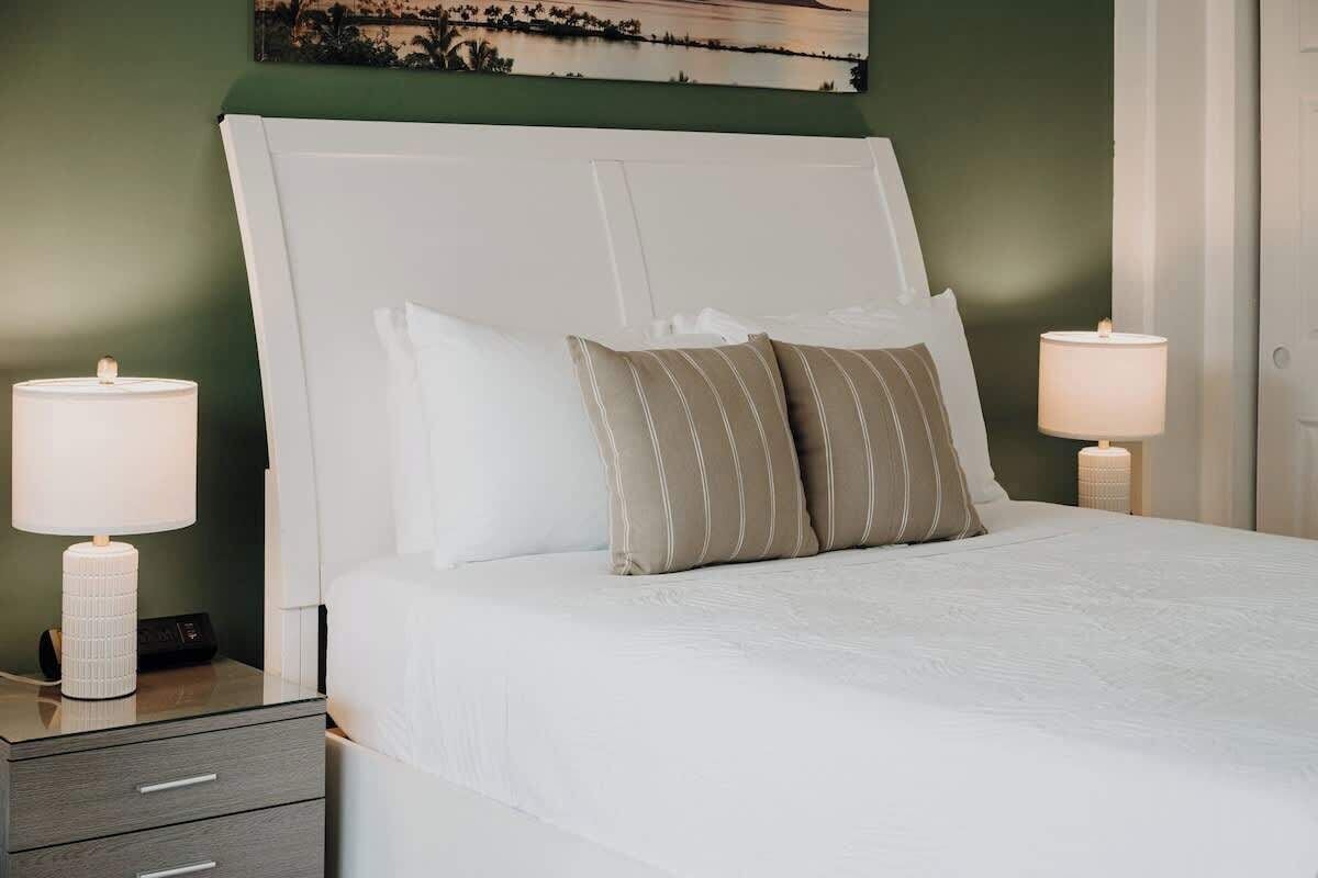 A neatly made bed with white bedding and striped pillows, a bedside table with a lamp, and a wall-mounted artwork above the bed.