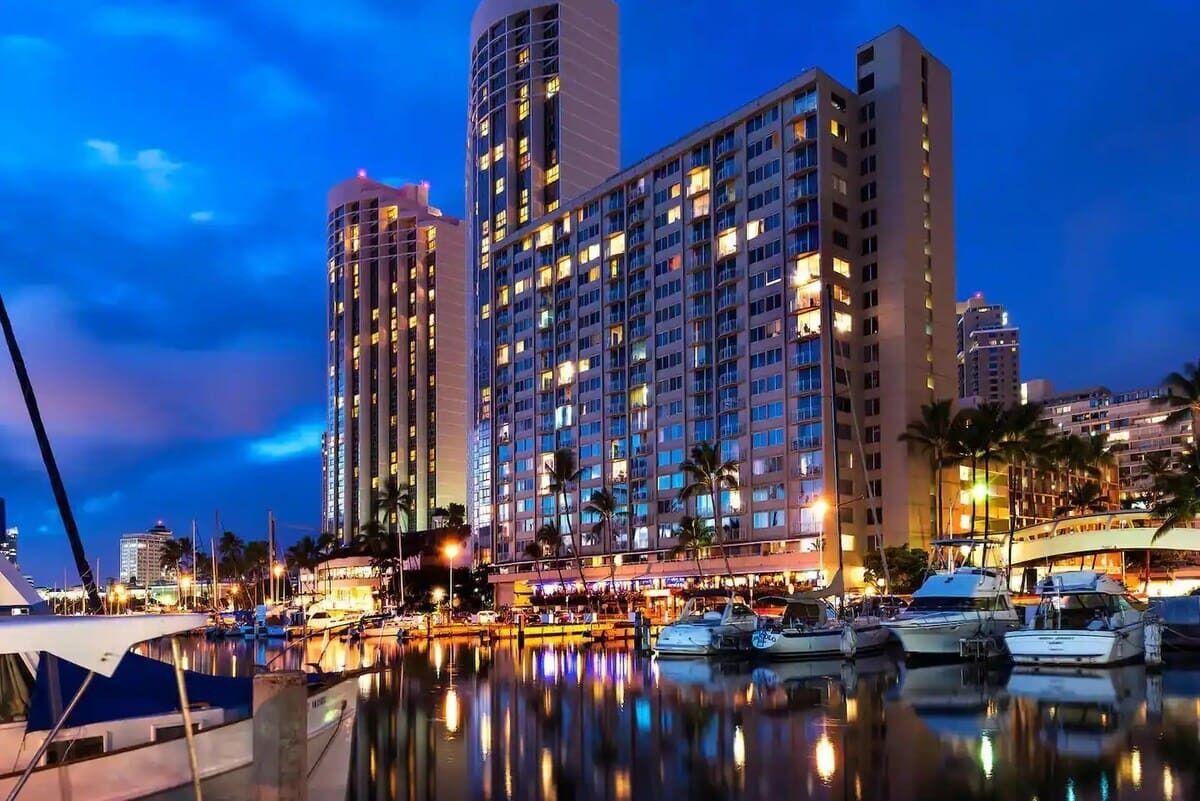 Una marina amb vaixells amarrats, un hotel gran de diversos pisos, i un cel serè.