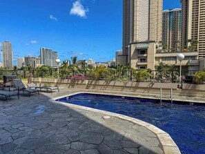 A swimming pool with lounge chairs, surrounded by a stone deck and palm trees, with tall buildings in the background. - Ocean View Studio, Newly Renovated, Free Parking (Honolulu)