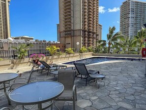 A rooftop pool area with lounge chairs and tables, surrounded by tall buildings and palm trees. - Ocean View Studio, Newly Renovated, Free Parking (Honolulu)