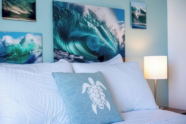 A bed with white linens, a blue pillow with a turtle design, and a wall with ocean wave paintings. - Ocean View Studio, Newly Renovated, Free Parking (Honolulu)