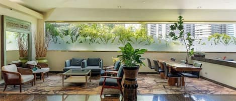 A spacious lobby with a large dining table, comfortable seating, and a view of greenery through frosted glass windows.