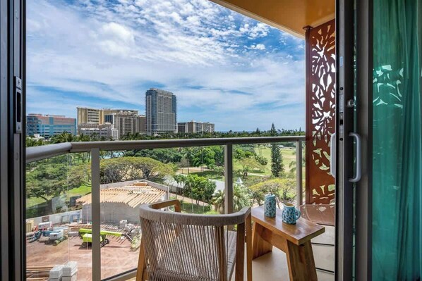 A balcony with a view of a cityscape, a wooden table, and a wicker chair. - Central Waikiki Studio, City View, Pool, 2 Queens (Honolulu)