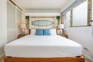 A bedroom with a large bed, two bedside tables, a wall-mounted artwork, and a window with blinds. - Central Waikiki 2 Bedroom, AC, Lanai, Pool, King (Honolulu)