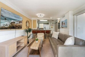 A living room with a sofa, a coffee table, a dining area with chairs, a kitchen in the background, and a large painting on the wall. - Central Waikiki 2 Bedroom, AC, Lanai, Pool, King (Honolulu)