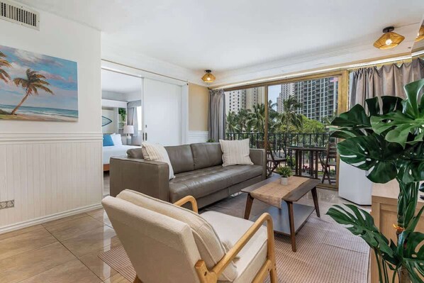 A modern living room with a sofa, chairs, a coffee table, and a large window with a view of palm trees and buildings. - Central Waikiki 2 Bedroom, AC, Lanai, Pool, King (Honolulu)