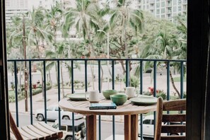 A balcony with a round table, chairs, and a view of palm trees and a street. - Central Waikiki 2 Bedroom, AC, Lanai, Pool, King (Honolulu)