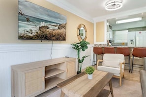 A living room with a large TV, a wooden cabinet, a plant, a white sofa, and a kitchen area in the background. - Central Waikiki 2 Bedroom, AC, Lanai, Pool, King (Honolulu)