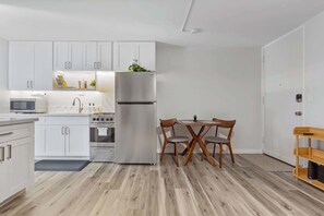 A modern kitchen with white cabinets, stainless steel appliances, a dining area with wooden chairs, and a small table. - Heart of Waikiki | Walkable, King Bed, Beach Gear. (Honolulu)