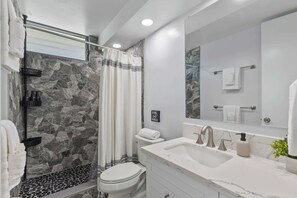 A modern bathroom with a stone wall, a shower with a glass door, a toilet, a sink with a marble countertop, and a mirror with a towel rack. - Heart of Waikiki | Walkable, King Bed, Beach Gear. (Honolulu)
