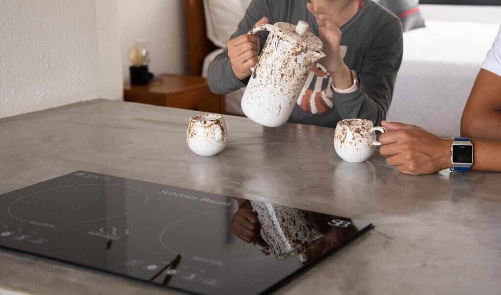 Une personne verse du café dans une tasse, une bouilloire et une tasse sont posés sur une table équipée d’une plaque de cuisson à induction numérique.