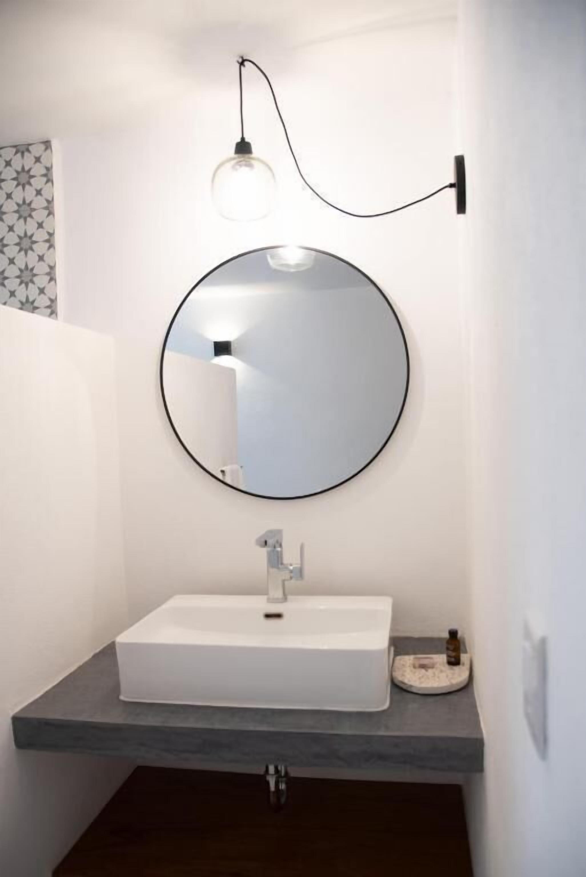 Une salle de bain avec un miroir rond, un lavabo blanc et un luminaire placé au-dessus du miroir.