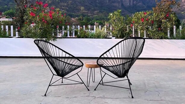 Una terraza con dos sillas de estructura metálica negra y una pequeña mesa redonda, con vistas a un paisaje montañoso con vegetación y flores rojas.