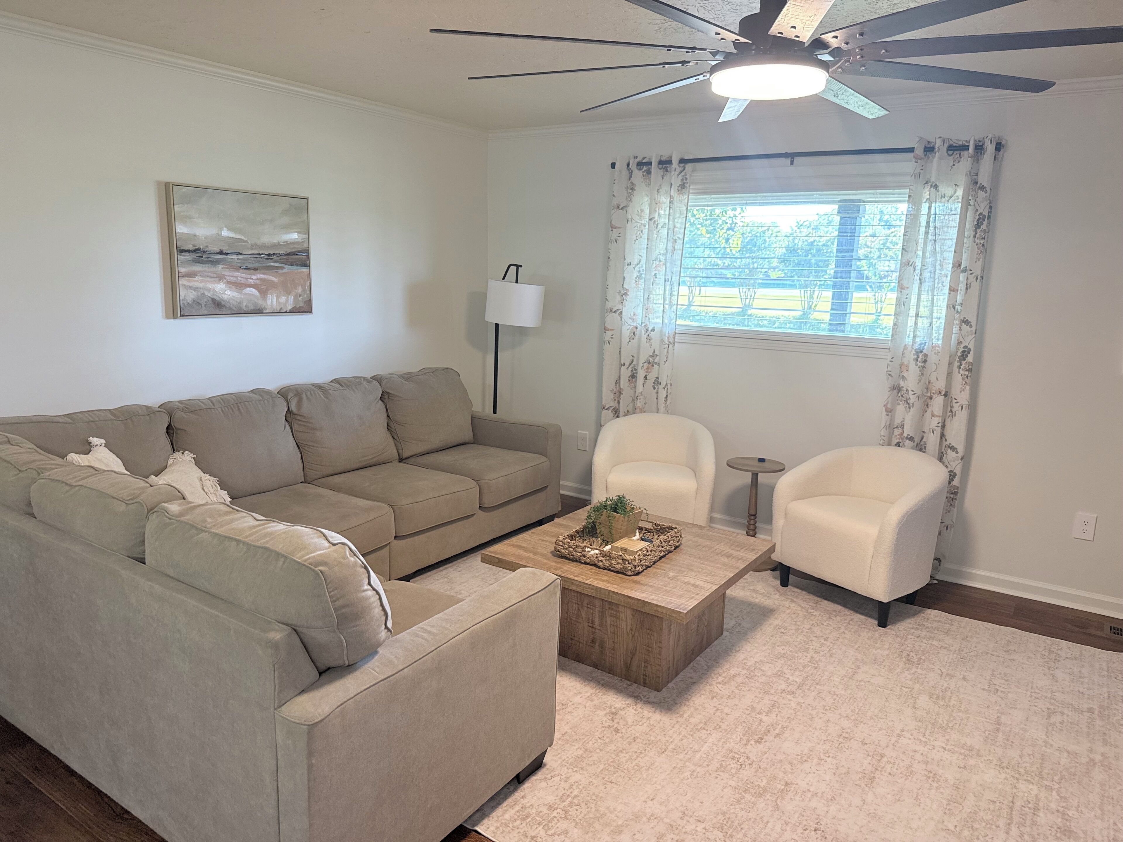 A living room with a sofa, two armchairs, a coffee table, a ceiling fan, a lamp, and a window with curtains.