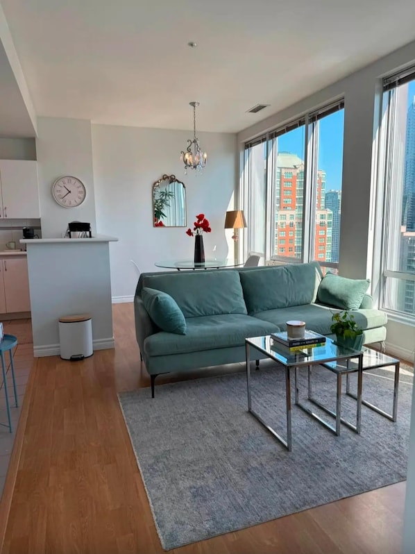 Un salon moderne avec un canapé, une table basse, un lustre, une horloge et une vue sur les bâtiments à travers de grandes fenêtres.