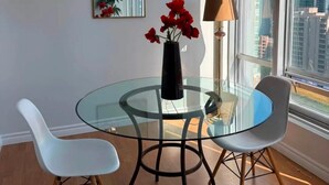 A dining area with a glass table, white chairs, a vase with red flowers, a chandelier, a mirror, and a view of the city through large windows.