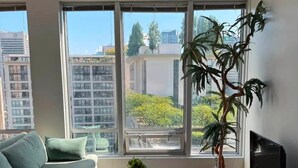 A modern living room with a sofa, a coffee table, a TV stand, and a large window with a view of buildings and trees.