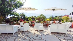 An outdoor seating area with white chairs and tables, surrounded by potted plants and umbrellas. - Xenia & Antonis  (Nikiana)