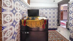 A room with patterned tile walls, a television on a stand, a small table with a book and remote, and a patterned rug. - Stay in the Heart of Marrakech Medina – Private Room, AC & Rooftop Terrace (Marrakech)