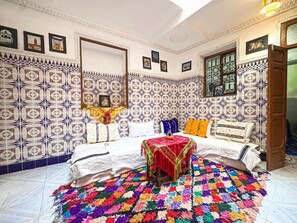 Une pièce avec un mur recouvert de carreaux à motifs, un lit avec du linge de lit blanc, un tapis coloré et une petite table recouverte d’une nappe rouge.