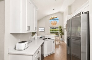 A modern kitchen with white cabinetry, a built-in island, a stainless steel refrigerator, and a window with a view of greenery. - Heated Pool~Game Room~Modern~Resort~Disney Magic (Kissimmee)