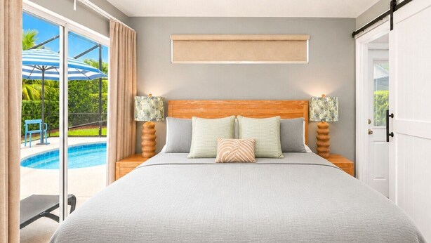 Un dormitorio con una cama grande, cabecera de madera, dos mesitas de noche con lámparas, vista a una piscina y una puerta corredera.