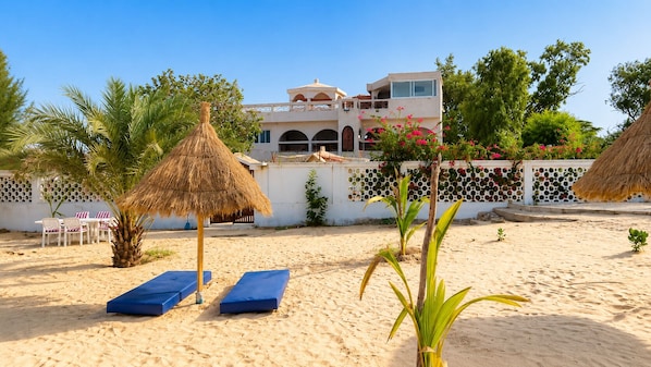 A beach with sun loungers, a thatched umbrella, and a building with a white facade and arches.