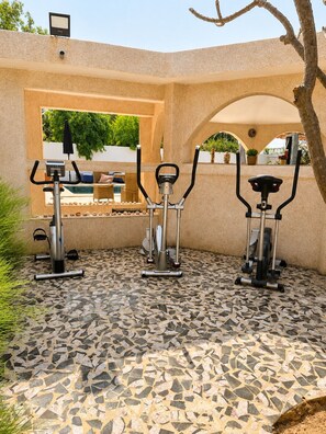Un espace extérieur aménagé pour faire de l’exercice, comprenant trois vélos d’appartement, un sol carrelé et une vue sur une piscine et un espace de détente.
