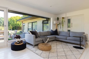 A modern living room with a grey sectional sofa, a round ottoman, a small side table, and a large window offering a view of the outdoors. - Scandi-style Birdham Retreat With Hot Tub Garden (Chichester)