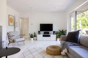 A modern living room with a flat-screen TV, a grey sofa, a round ottoman, and a white cabinet with speakers. - Scandi-style Birdham Retreat With Hot Tub Garden (Chichester)