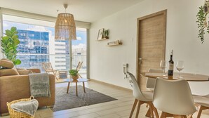 Una sala moderna con sofá, mesa de comedor, sillas, una lámpara colgante y vistas de un paisaje urbano a través de amplios ventanales.