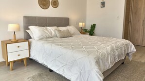 Un dormitorio con cama, mesitas de noche, una lámpara de pared, una planta en maceta y una puerta.