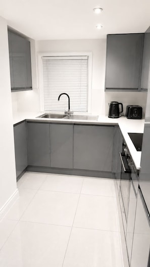 Eine moderne Küche mit grauen Schränken, einer Edelstahl-Spüle, einem Fenster mit Jalousien, einem Wasserkocher und einem Toaster.