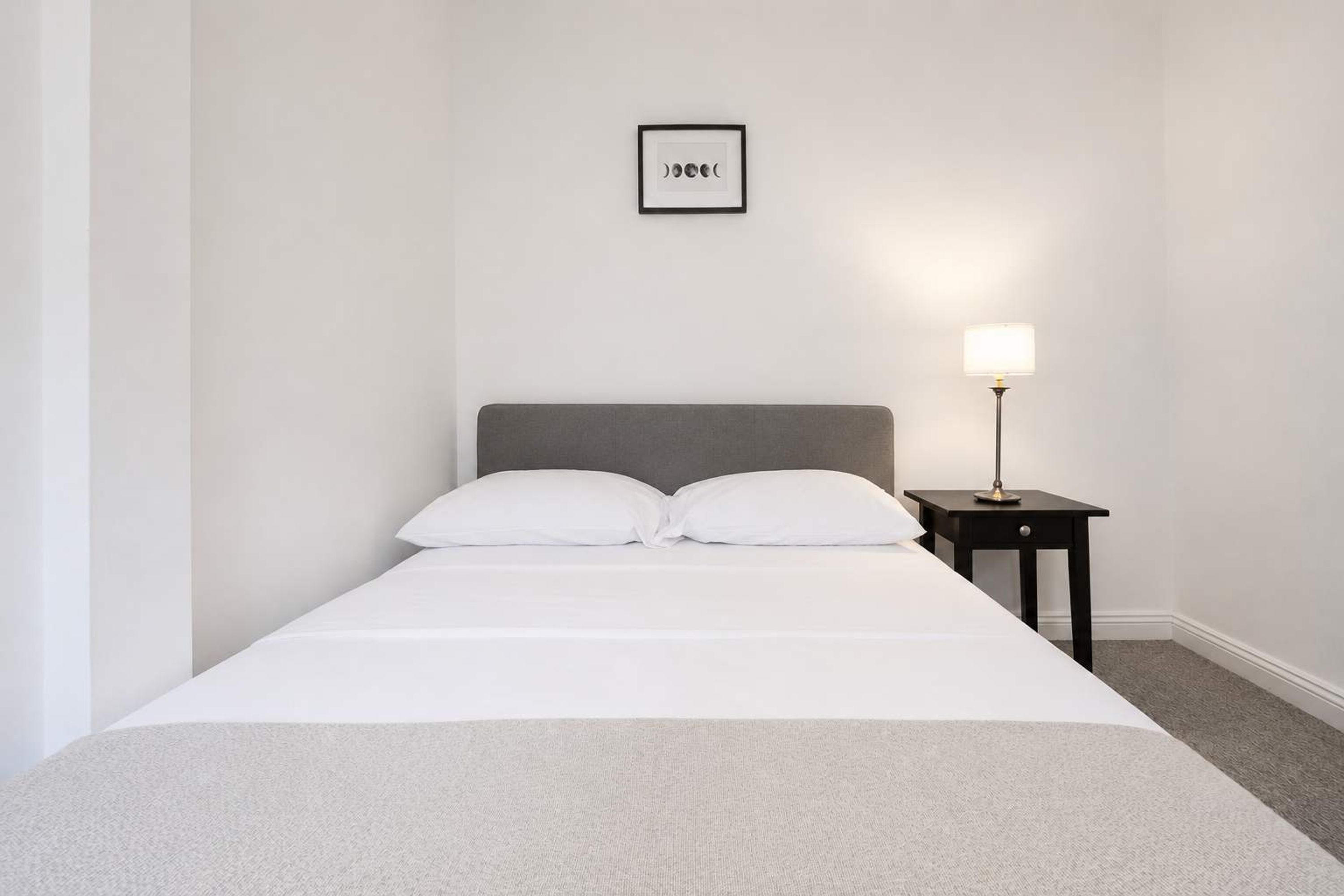 A neatly made bed with white bedding, a grey headboard, and a small bedside table with a lamp.
