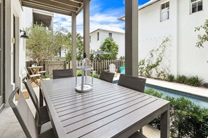 Um alpendre coberto com mesa e cadeiras, um candeeiro, uma piscina e um edifício branco ao fundo.