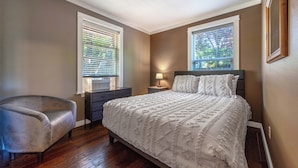 Quarto com cama, cadeira, cômoda e duas janelas com persianas.