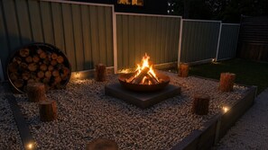 Ein Lagerfeuer, umgeben von Kies, mit Holzscheiten in der Nähe und Baumstümpfen darumherum.