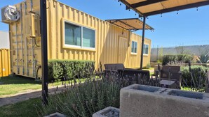 En gul, containerlignende bygning med terrasse, udendørs møbler og en have med lavendelplanter.