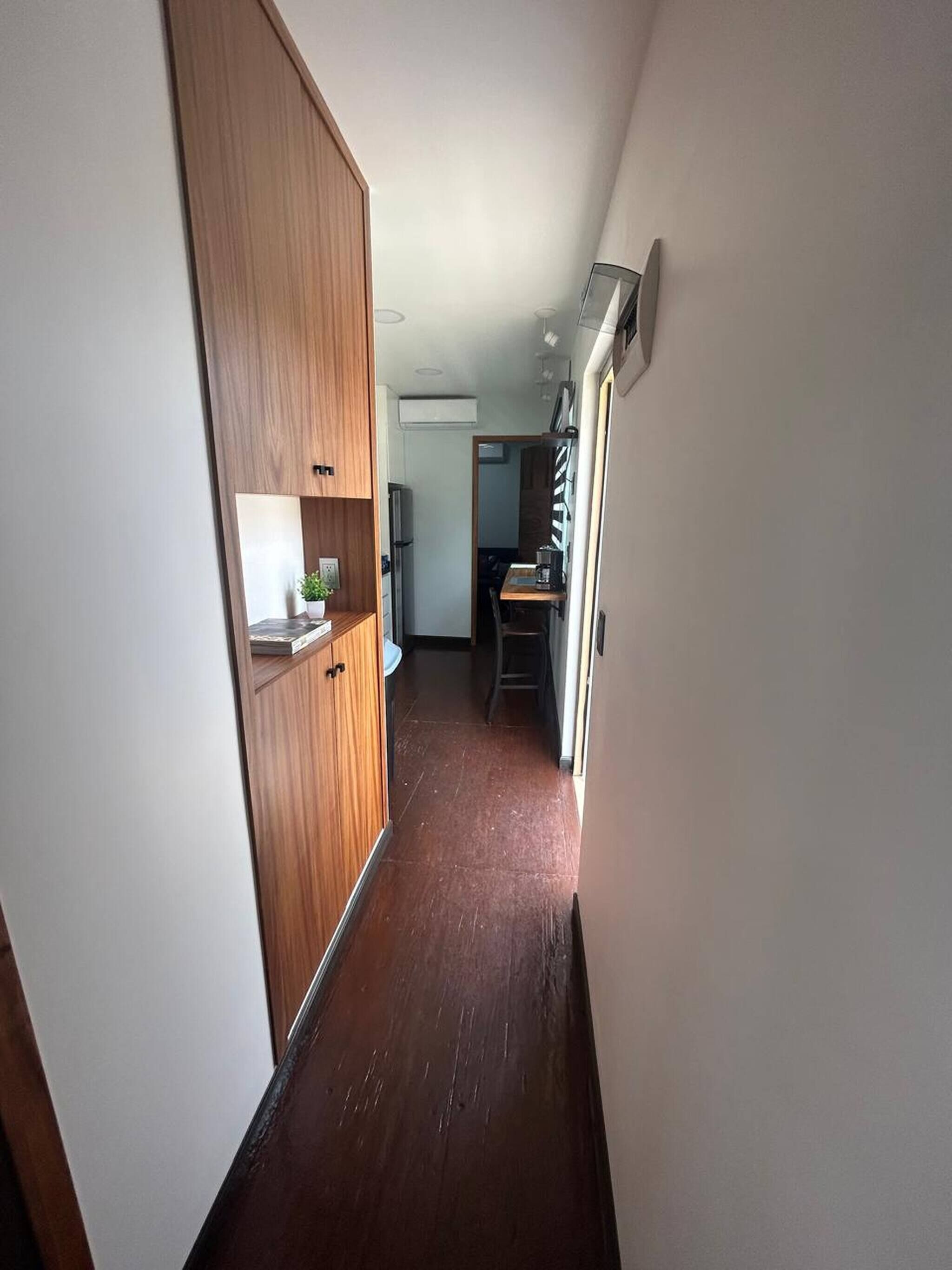 A narrow hallway with wooden cabinets, a kitchen area with a refrigerator, and a dining table.