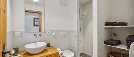 Un baño con tocador de madera, lavamanos blanco, inodoro, ducha y estantes con toallas.