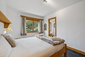 Ein Schlafzimmer mit Bett, einem Vorhangfenster, einer Lampe, einem Spiegel und Blick auf Grünfläche.
