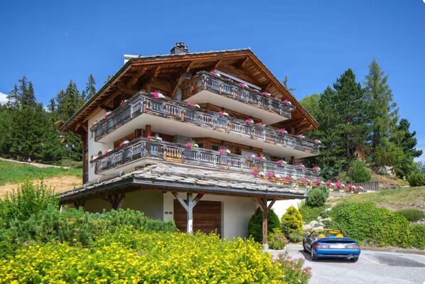 Ein mehrstöckiges Chalet mit Balkonen, umgeben von Grün und einem parkenden Auto.