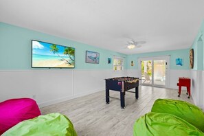 A room with a foosball table, a TV with a beach scene, and a sliding glass door leading to an outdoor area. - Sunset at 714 - Luxury Island Retreat with Heated Pool Near Bean Point Beach (Anna Maria)