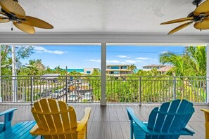 A balcony with two Adirondack chairs, a ceiling fan, and a view of a resort area with buildings and palm trees. - Sunset at 714 - Luxury Island Retreat with Heated Pool Near Bean Point Beach (Anna Maria)