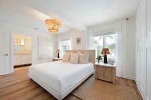 A spacious bedroom with a large bed, a bedside table with a lamp, a nightstand, and a view of the outdoors through a window. - The Palm at Bean Point - Luxury Coastal Retreat with Heated Pool, Walk to Beach (Anna Maria)