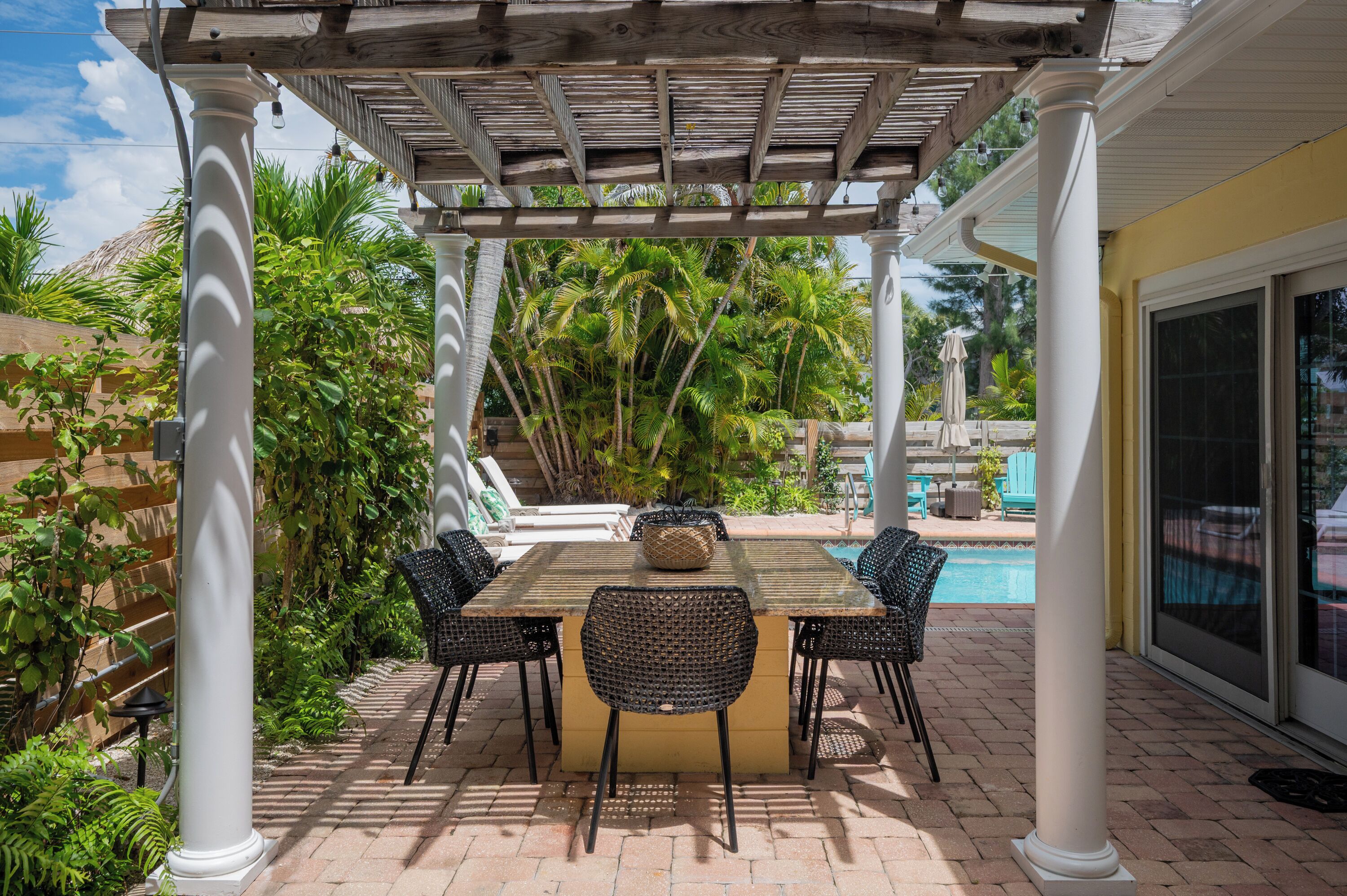 A covered patio with a table and chairs, surrounded by lush greenery and a pool.