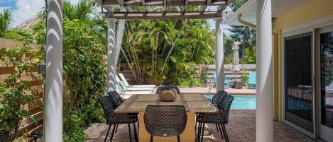 A covered patio with a table and chairs, surrounded by lush greenery and a pool.
