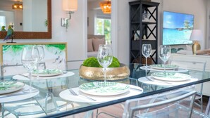 Zona pranzo con tavolo in vetro apparecchiato per quattro persone, una ciotola decorativa verde e un televisore che mostra una spiaggia.