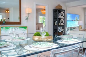 A dining area with a glass table set for four, a green decorative bowl, and a television displaying a beach scene. - The Palm at Bean Point - Luxury Coastal Retreat with Heated Pool, Walk to Beach (Anna Maria)